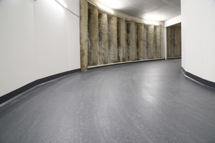 Tiefgarage mit einem gewundenen, grauen Boden und einer Wand, die aus vertikalen, strukturierten Elementen besteht. Die Wände sind weiß und die Beleuchtung ist gleichmäßig.