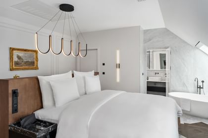 Modernes Schlafzimmer der Loft Suite im „The Amauris“ Wien mit großem Doppelbett, Holz-Kopfteil, organischer schwarzer Deckenleuchte, Wandgemälde und Blick in ein helles Badezimmer mit freistehender Badewanne und Marmorflächen.