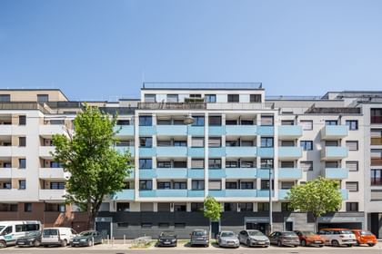 Straßenansicht der Fassade eines Mehrparteienhauses, blaue Balkone, davor parken Autos