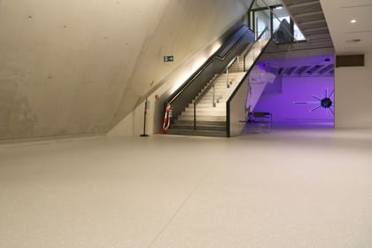 Moderner Ausstellungsraum mit weißen Wänden und Bodenbeschichtung im Schlossmuseum Linz. eine Treppe mit schwarzem Glasgeländer führt hinauf zum Ausgang