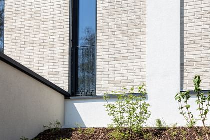  Nahaufnahme einer Gebäudefassade mit hellen Ziegeln, schwarzen Fensterbänken und Metallgittern an den Fenstern. Unten und rechts ist weißer Putz zu sehen, entlang der Wand verläuft ein Streifen mit weißen Kieselsteinen.