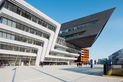 Außenansicht der Wirtschaftsuniversität Wien mit moderner, weißer Fassade, großen Glasflächen und geometrischen Formen vor blauem Himmel.