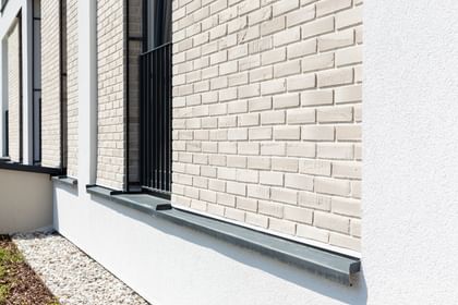 Nahaufnahme einer Gebäudefassade mit hellen Ziegeln, schwarzen Fensterbänken und Metallgittern an den Fenstern. Unten und rechts ist weißer Putz zu sehen, entlang der Wand verläuft ein Streifen mit weißen Kieselsteinen.