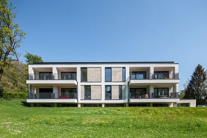 Modernes zweistöckiges Gebäude mit großen Fenstern und Balkonen, gelegen in einer grünen Umgebung mit Bäumen im Hintergrund bei sonnigem Wetter und klarem Himmel.