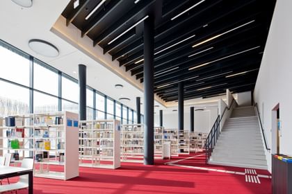 Moderner, heller Bibliotheksraum der Wirtschaftsuniversität Wien mit rotem Teppich, weißen Bücherregalen, großen Glasfronten und schwarzen Säulen. Eine breite Treppe führt ins offene, lichtdurchflutete Obergeschoss.