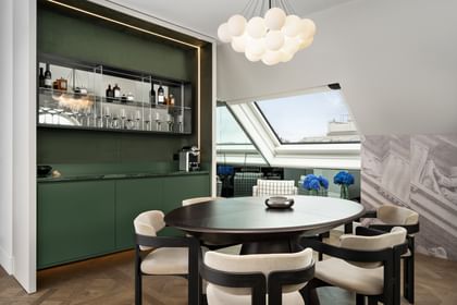 Ess- und Loungebereich der Loft Suite im „The Amauris“ Wien mit rundem Holztisch und vier gepolsterten Stühlen, eleganter grüner Einbau-Bar mit Spiegelregal, Flaschen und Gläsern sowie großen Dachfenstern mit Stadtblick und blauen Hortensien.