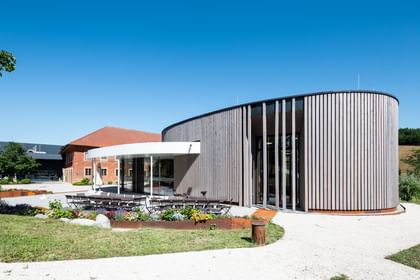 Gebäude mit einer geschwungenen Holzfassade und großen Fenstern, umgeben von einem gepflegten Garten und einer Terrasse