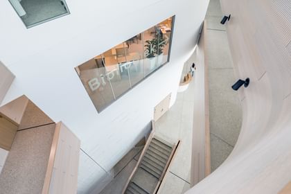 Innenansicht der Anton Bruckner Privatuniversität mit zentraler Treppe, weißen Wänden und Holzakzenten. Auf einer oberen Ebene befindet sich ein Fenster mit der Aufschrift „Bibliothek“.