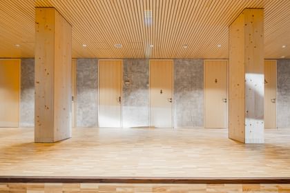 Innenraum mit einem hellen Holzboden und Wänden in Betonoptik. In der Mitte des Raumes stehen mehrere Säulen, die den Raum strukturieren. An der Wand sind mehrere Türen, die in weitere Räume führen.