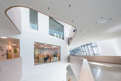 Innenansicht der Anton Bruckner Privatuniversität mit geschwungener Architektur und großem Fenster mit der Aufschrift „Bibliothek“. Helle, lichtdurchflutete Umgebung mit perforierter Decke, Lichtinstallationen und Wandpostern.