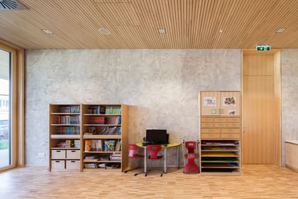 Lern- oder Arbeitsbereich einer Schule. Auf der linken Seite befindet sich ein Bücherregal mit verschiedenen Büchern und Spielmaterialien. In der Mitte steht ein kleiner Tisch mit zwei roten Stühlen, an dem ein schwarzer Computer platziert ist.