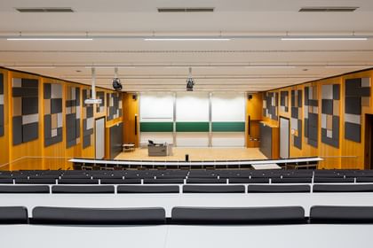 Leerer Hörsaal mit gestuften Sitzreihen und langen Tischen, grüner Tafel, doppelflügeliger Tür und Uhr. Die Wände sind mit Akustikpaneelen in verschiedenen Formen versehen, die Decke mit mehreren Lichtquellen ausgestattet.