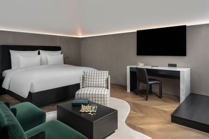 Eleganter Wohn- und Schlafbereich in der Grand-Maisonette-Suite des „The Amauris“ Wien mit großem Doppelbett, Schreibtisch mit TV, zwei dunkelgrünen Sesseln, schwarzem Couchtisch mit Schachbrett und kariertem Sessel auf hellem Teppich und Fischgrätparkett.