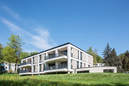 Modernes zweistöckiges Gebäude mit großen Fenstern und Balkonen, gelegen in einer grünen Umgebung mit Bäumen im Hintergrund bei sonnigem Wetter und klarem Himmel.