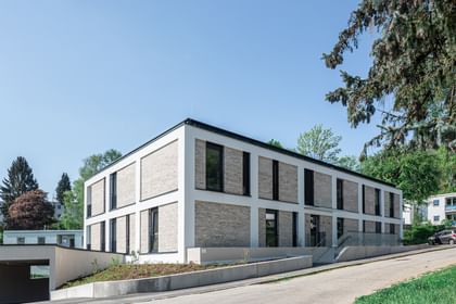Modernes zweistöckiges Gebäude mit großen Fenstern und Balkonen, gelegen in einer grünen Umgebung mit Bäumen im Hintergrund bei sonnigem Wetter und klarem Himmel.