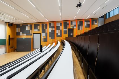 Leerer Hörsaal mit gestuften Sitzreihen und langen Tischen, grüner Tafel, doppelflügeliger Tür und Uhr. Die Wände sind mit Akustikpaneelen in verschiedenen Formen versehen, die Decke mit mehreren Lichtquellen ausgestattet.