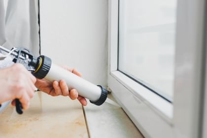 Eine Person verwendet eine Silikonpistole, um Dichtmasse entlang des Rahmens eines Fensters aufzutragen.