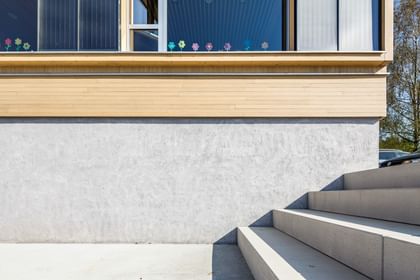 Außenbereich eines Gebäudes mit einer hellen, strukturierten Wand und einer Treppe, die zu einem Eingang führt. Im oberen Bereich des Bildes sind bunte Blumenaufkleber an einem Fenster angebracht