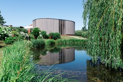 Rundes Gebäude mit einer Holzfassade spiegelt sich in einem ruhigen Teich, umgeben von üppigem Grün und blühenden Pflanzen