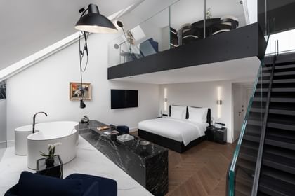 Zweistöckige Grand-Maisonette-Suite im „The Amauris“ Wien mit freistehender weißer Designerbadewanne auf Marmor, Doppelbett mit weißer Bettwäsche, wandmontiertem Flachbildfernseher und schwarzer Treppe mit Glasgeländer zu einer offenen Galerie mit Sitzecke.