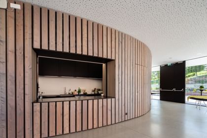 Minimalistische Küche oder Bar in einem Raum mit einer geschwungenen Wand aus Holzlatten. Die Wand ist hell und hat eine natürliche Holzfarbe. Im Hintergrund ist eine dunkle Küchenzeile sichtbar, die mit verschiedenen Getränken und Utensilien ausgestattet ist