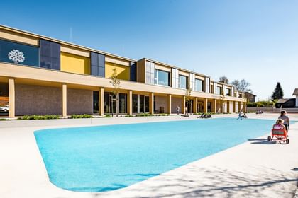  Außenansicht der Volkshochschule. Im Vordergrund sind zwei Kinder auf einem roten Spielzeugfahrzeug zu sehen, die vor einer blauen Spielfläche spielen, während andere Kinder mehr im Hintergrund spielen