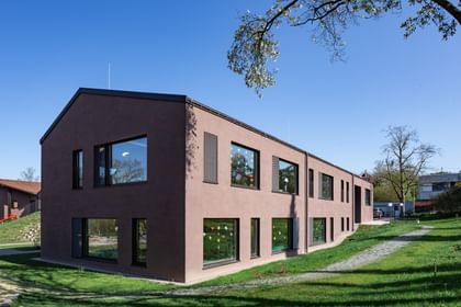 Zweigeschossiges Gebäude mit großen Fenstern, das im Grünen steht. In den Fenstern sind bunte Zeichnungen sichtbar.