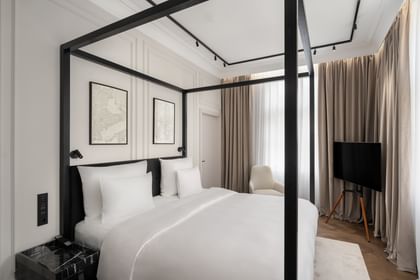 Helles Executive-Suite-Schlafzimmer im „The Amauris“ Wien mit modernem Himmelbett in schwarzem Rahmen, weißer Bettwäsche, abstrakten Wandbildern, bodentiefen Vorhängen, Lounge-Sessel und freistehendem TV auf hellem Parkett.