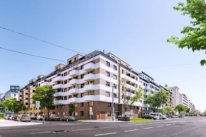 Außenansicht eines mehrstöckigen Gebäudes mit Balkonen. Im Vordergrund verläuft eine Straße und vor dem Gebäude parken mehrere Autos.