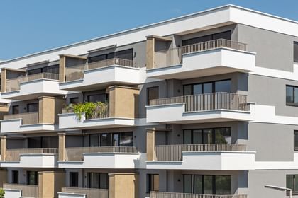 Wohnanlage in der Robert-Stolz-Straße in Linz. Ein modernes, mehrstöckiges Wohngebäude mit hellen Fassaden und zahlreichen Balkonen. Große Fenster sorgen für viel Tageslicht. Vor dem Gebäude stehen Autos auf einer ruhig wirkenden Straße, eingerahmt von gepflegten Grünflächen und Bäumen.