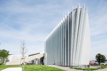 Modernes Gebäude der Anton Bruckner Privatuniversität mit geschwungener Fassade und vertikalen weißen Lamellen an der Außenwand. Umgeben von Grünflächen und Bäumen bei sonnigem Wetter mit leicht bewölktem Himmel.