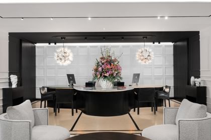 Helle Hotellobby des „The Amauris“ Wien mit halbkreisförmiger dunkler Rezeptionstheke, großem Blumenarrangement, drei Arbeitsplätzen vor kassettierten weißen Wänden, zwei kugelförmigen Glasleuchten und zwei gepolsterten Lounge-Sesseln im Vordergrund.