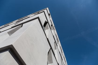 Die Ecke eines Gebäudes, die in die Höhe ragt. Die Fassade ist aus hellem Beton mit schmalen Fenstern, vor einem klaren blauen Himmel.