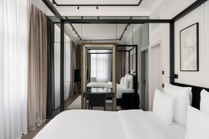 Helles Executive-Suite-Zimmer im „The Amauris“ Wien mit großem Doppelbett, weißer Bettwäsche, Spiegelfront mit Schreibtisch und schwarzem Stuhl, bodentiefen Vorhängen und gemütlicher Sitzecke mit Sessel und TV im Hintergrund.