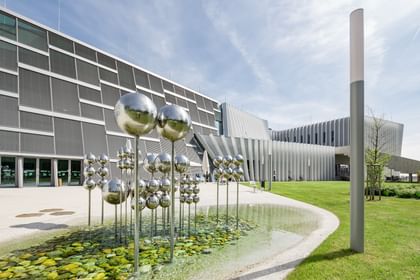 Modernes Gebäude der Anton Bruckner Privatuniversität mit markanter Linienarchitektur und großen Fenstern. Im Vordergrund eine künstlerische Installation aus metallischen Kugeln auf Stangen, platziert in einem flachen Wasserbecken mit grünen Steinen.