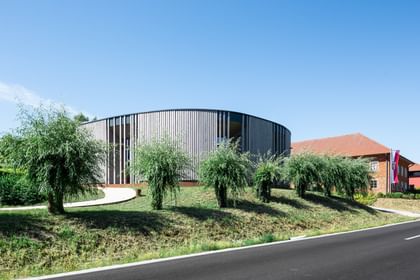 Rundes Gebäude mit einer Holzfassade, das von einer grünen Landschaft umgeben ist. Im Vordergrund sind mehrere Sträucher oder kleine Bäume zu sehen