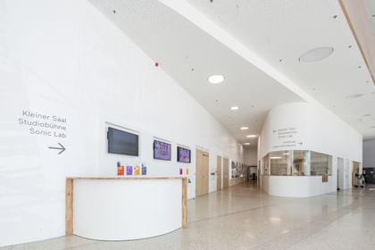 Moderner, heller Flur in der Anton Bruckner Privatuniversität mit weißen Wänden und poliertem Boden. Wegweiser zu „Kleiner Saal“, „Studiobühne“, „Sonic Lab“ und „Unterrichtsräume“ sowie drei Wandbildschirme und ein Informationscounter mit Holzplatte und Flyern.