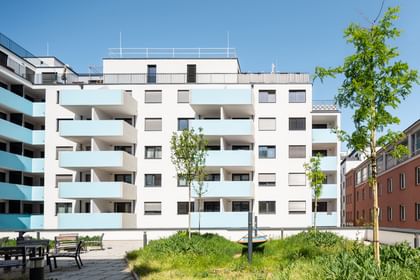 Mehrstöckiges Wohnhaus mit weißer Fassade, türkisen Balkonen, Rasen und Bäumen
