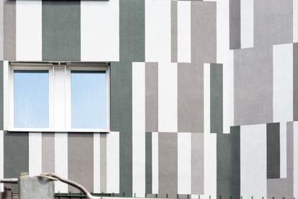 Ein Gebäude mit einer Fassade, die aus vertikalen und horizontalen Streifen in verschiedenen Grautönen und Weiß besteht. In der Mitte der Wand befindet sich ein Fenster mit zwei Fensterscheiben.