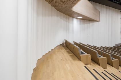 Detailansicht eines modernen Konzert- oder Vortragssaals der Anton Bruckner Privatuniversität mit wellenförmiger, weißer Akustikwand, hellem Holzboden und -decke sowie gepolsterten Holzsitzen; das Design betont hochwertige Materialien, optimale Akustik und eine elegante, zeitgemäße Raumgestaltung.