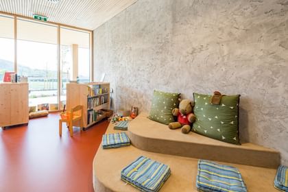 Im Vordergrund befinden sich zwei große, grüne Kissen auf einer erhöhten Sitzfläche aus hellem Holz. Daneben liegen gestreifte Decken. Im Hintergrund steht ein Bücherregal mit einer Auswahl an Kinderbüchern. Auf dem Boden sind einige Spielzeuge und ein Teddybär zu sehen. 