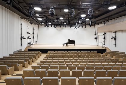 Blick von hinten in einen modernen Konzertsaal der Anton Bruckner Privatuniversität mit ansteigenden Sitzreihen, offen gestalteter Bühne und Flügel; Beleuchtung und klar strukturierte Architektur sorgen für ein helles, freundliches Ambiente.