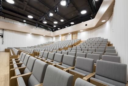 Moderner, großzügiger Hörsaal der Anton Bruckner Privatuniversität mit ansteigenden, gepolsterten Holzsitzreihen und hell gestalteter, wellenförmiger Wandverkleidung; ausgestattet mit moderner Deckenbeleuchtung und Veranstaltungstechnik – ideal für Vorträge, Konferenzen oder Präsentationen.