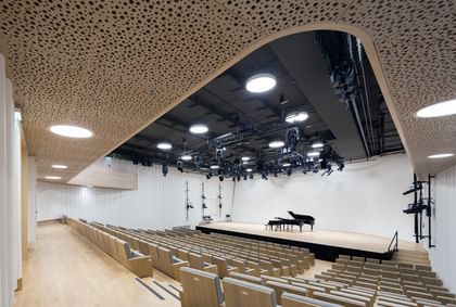 Blick von hinten in einen modernen Konzertsaal der Anton Bruckner Privatuniversität mit ansteigenden Sitzreihen, offen gestalteter Bühne und Flügel; hochwertige Akustikdecken, dezente Beleuchtung und klar strukturierte Architektur sorgen für ein helles, freundliches Ambiente.