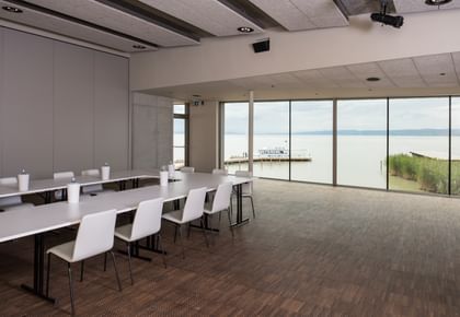 Konferenzsaal mit einem U-förmigen weißen Tisch und davorstehenden Stühlen und Ausblick auf den See