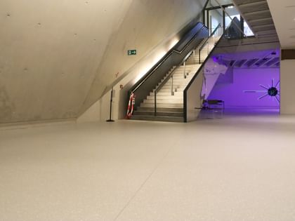 Moderner Ausstellungsraum mit weißen Wänden und Bodenbeschichtung im Schlossmuseum Linz. eine Treppe mit schwarzem Glasgeländer führt hinauf zum Ausgang