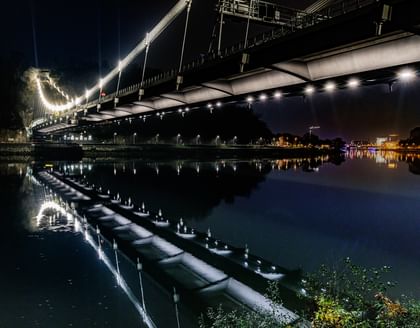 Eine nächtliche Luftaufnahme einer Brücke, die über einen Fluss führt. Die Brücke ist beleuchtet und reflektiert im Wasser.