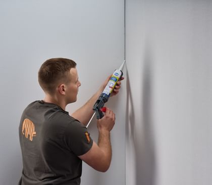 Ein Handwerker, der eine Dichtmasse in die Ecke einer Wand aufträgt. Er trägt ein graues T-Shirt und hält eine Kartusche mit Dichtmittel in der Hand.