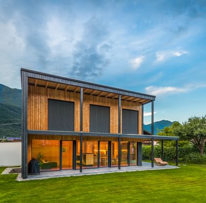Modernes zweistöckiges Haus mit großen Fenstern und Holzfassade, in der Abenddämmerung beleuchtet. Das Haus ist von grünem Rasen und einem gepflegten Garten umgeben, im Hintergrund sind Berge und weitere Häuser zu sehen.