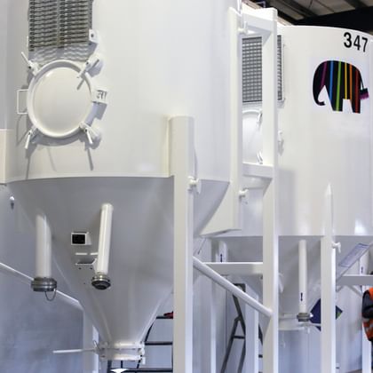 Mehrere große, weiße Silos mit Caparol-Logo und Nummerierung stehen nebeneinander in einer Industriehalle. Eine Person in Warnweste arbeitet im Hintergrund an einem der Silos.